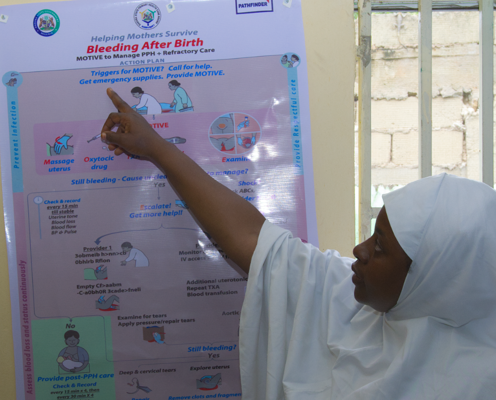 Health workers in Kaduna stand beside E-MOTIVE guidance displayed on a maternity ward wall as a reminder for managing postpartum haemorrhage